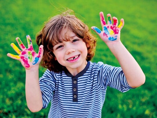 boy with curly hair without front milk tooth shows hands dirty with multi-colored finger paints and smiles.