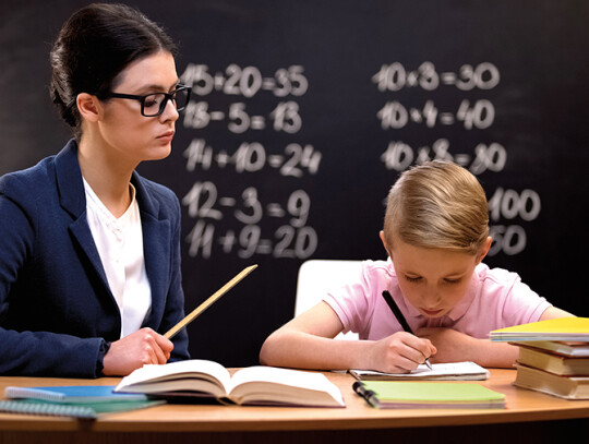 Strict teacher with ruler watching schoolboy doing his homework,